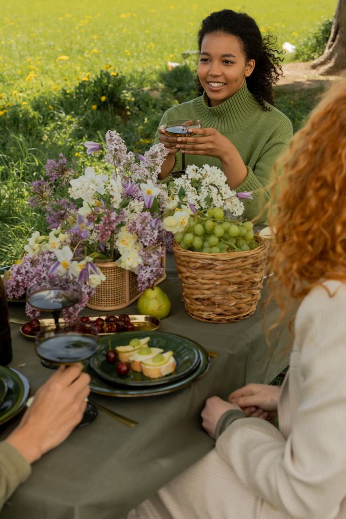 Friends enjoying a picnic outdoors with flowers, fruits, and wine in a lush, sunny setting.